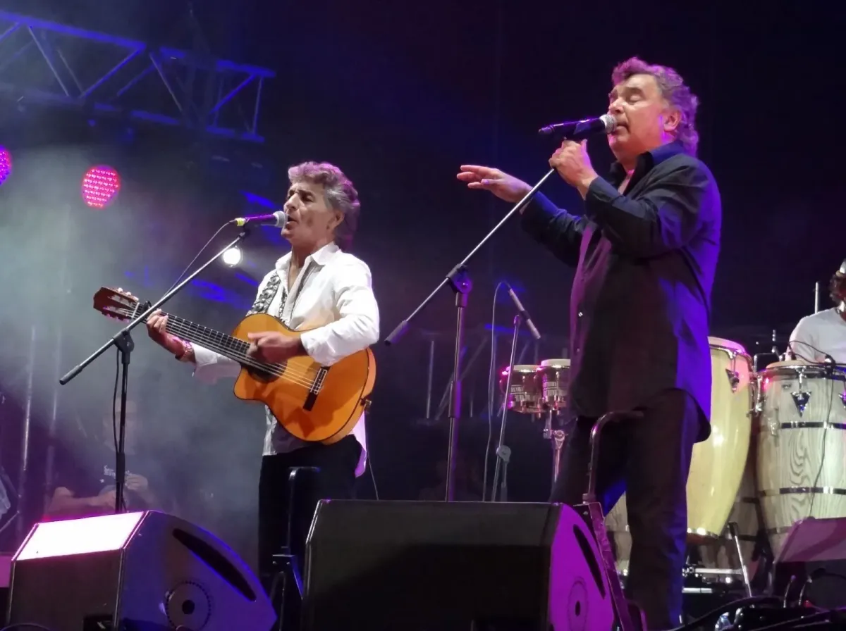 Dois integrantes do Gipsy Kings cantam e tocam violão em palco com iluminação roxa