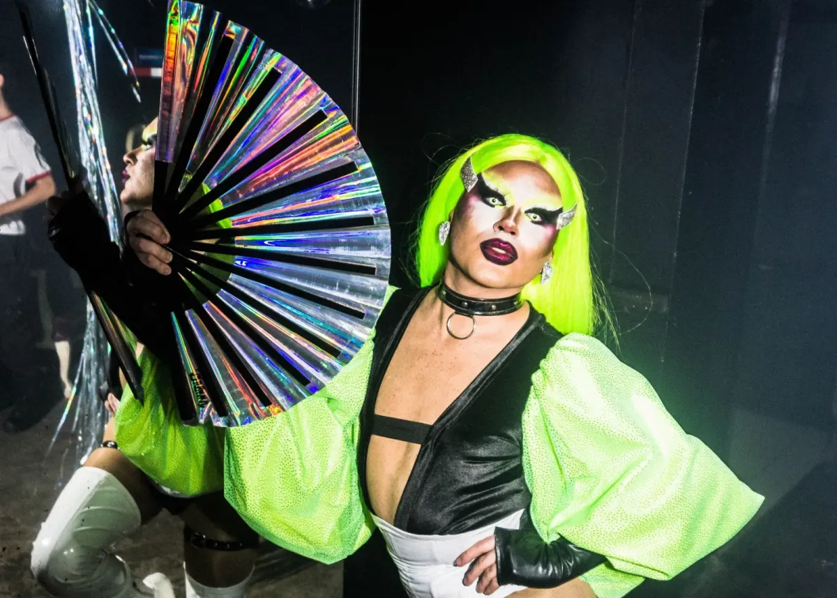 Artista drag com cabelo verde neon e leque holográfico em palco escuro