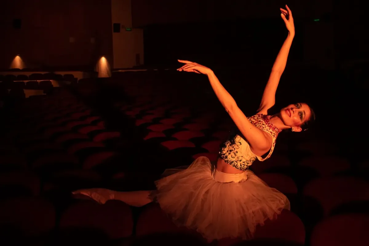 Bailarina de tutu com os braços erguidos, iluminada por luz quente, em um teatro com poltronas vermelhas