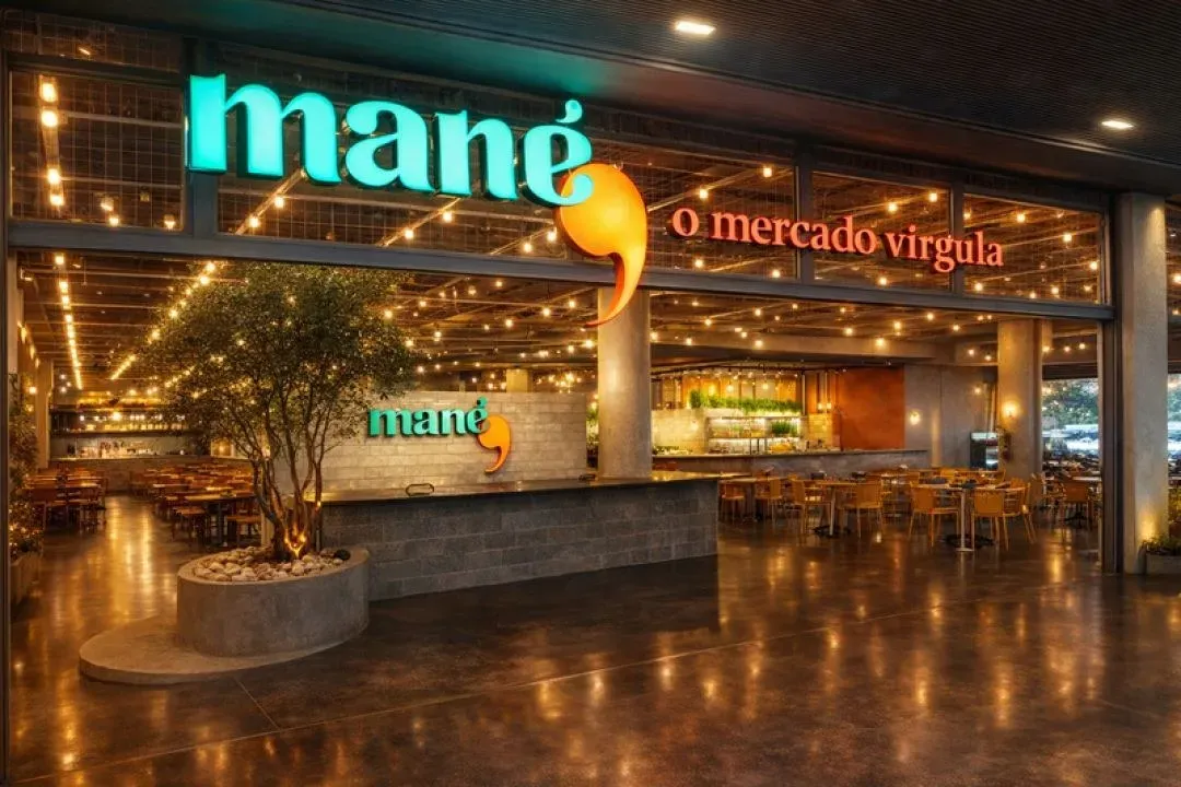 Salão iluminado de uma unidade do Mané Mercado, com letreiro em neon verde, árvore no centro e mesas