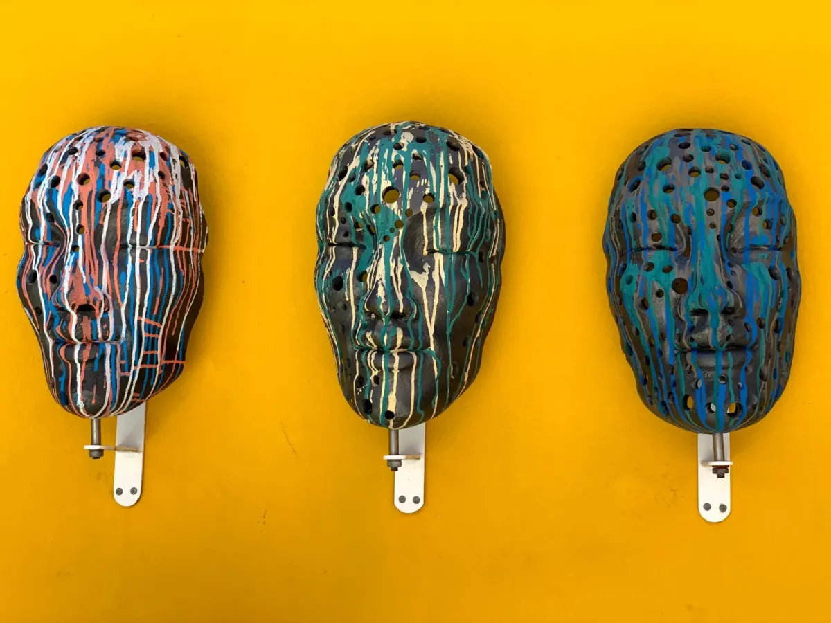 Três máscaras de cerâmica escura com respingos de tinta colorida, presas a uma parede amarela; foto ilustrativa, não são obras da exposição