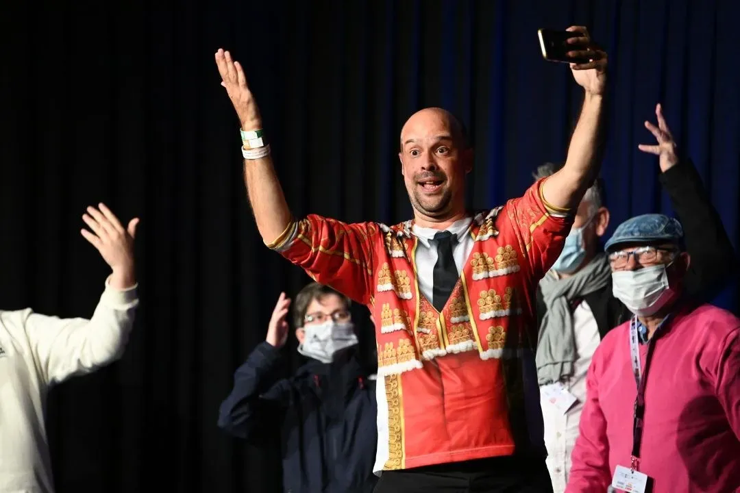 Artista surdo de casaco vermelho ergue os braços no palco, com o público fazendo sinais em Libras