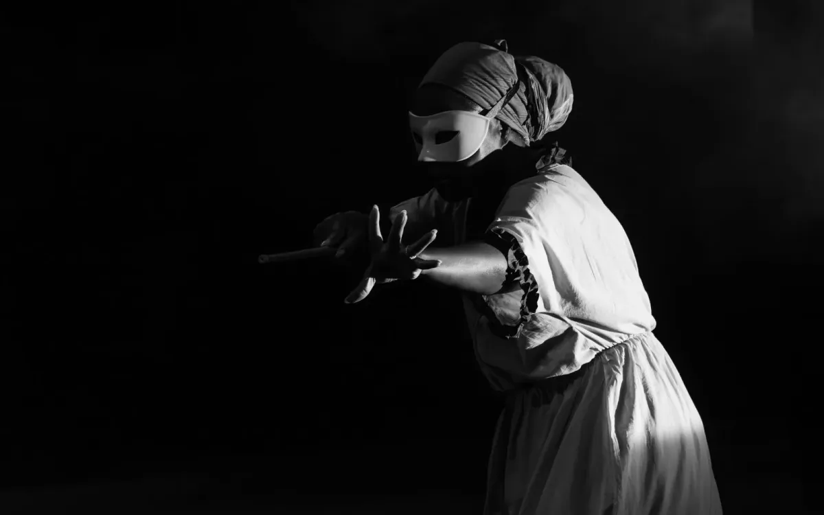 Artista de máscara branca e figurino claro em cena, com luz lateral sobre fundo escuro (foto ilustrativa de teatro)