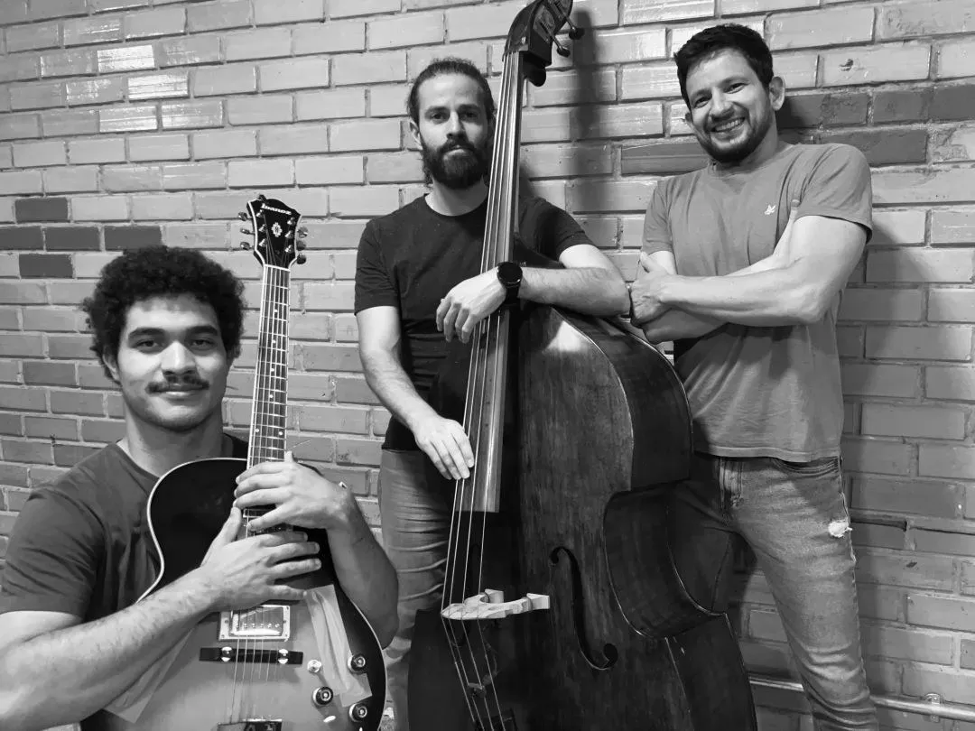 Os três músicos da banda Dona Sarah du Jazz, com guitarra e contrabaixo acústico, encostados numa parede de tijolos