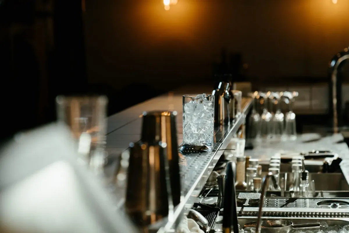 Balcão de bar com copos, coqueteleiras e gelo, em ambiente de luz baixa