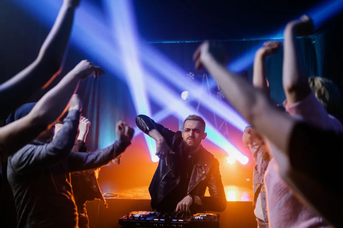 DJ na cabine com luzes azuis e laranja e plateia de braços erguidos, imagem ilustrativa de festa eletrônica