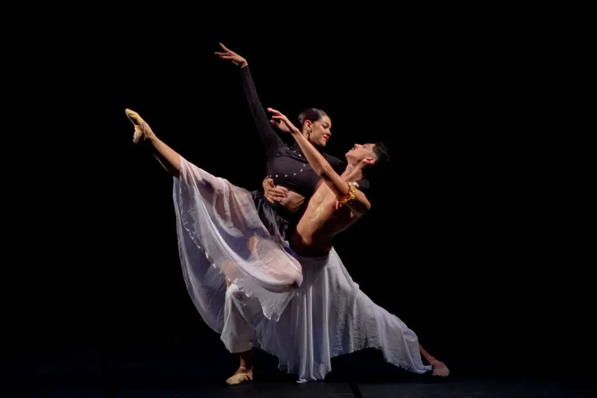 Casal de bailarinos em passo a dois, de figurino claro, sobre um palco escuro