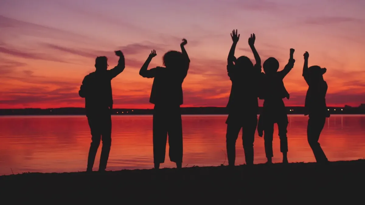 Silhueta de um grupo de pessoas dançando de braços erguidos à beira de um lago ao pôr do sol