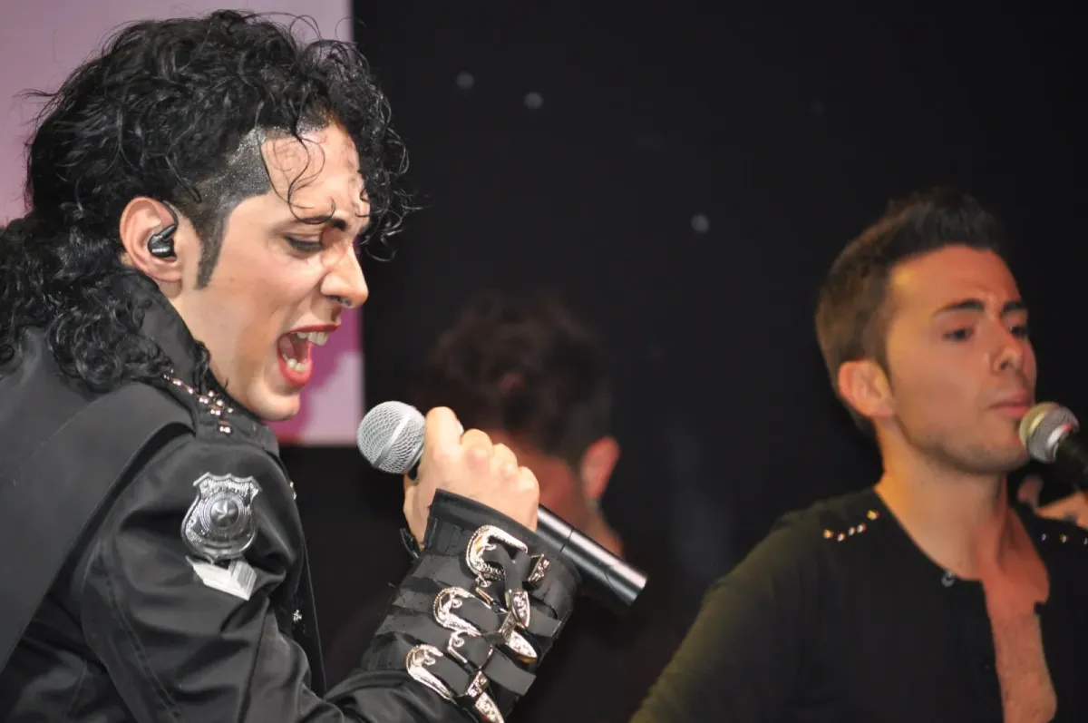 Artista de jaqueta preta com fivelas prateadas canta ao microfone em show tributo a Michael Jackson