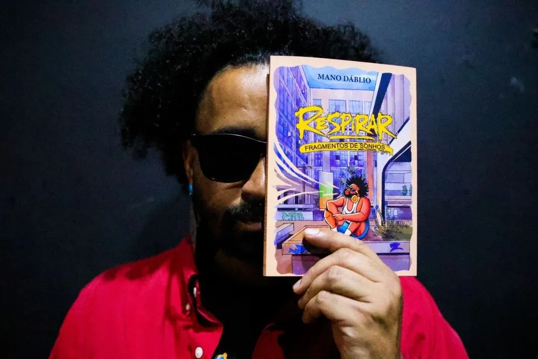 O poeta Mano Dáblio, de óculos escuros e camisa vermelha, segura o livro Respirar em frente ao rosto