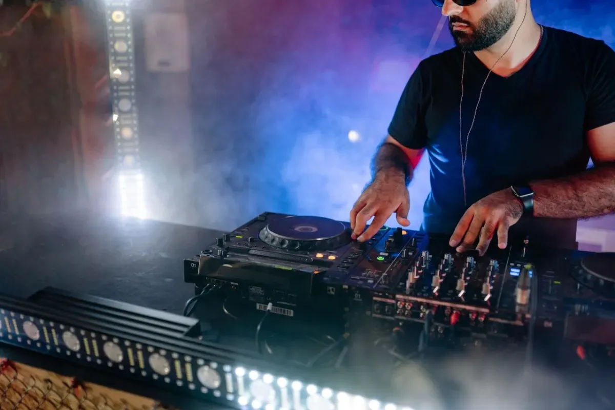 DJ de camiseta preta diante de equipamento de mixagem, com fumaça e luzes no palco, em foto ilustrativa