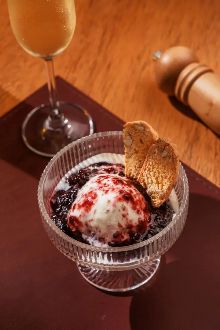 Taça de gelato de fior di latte com calda de frutas vermelhas e biscoito cantucci, ao lado de uma taça de espumante