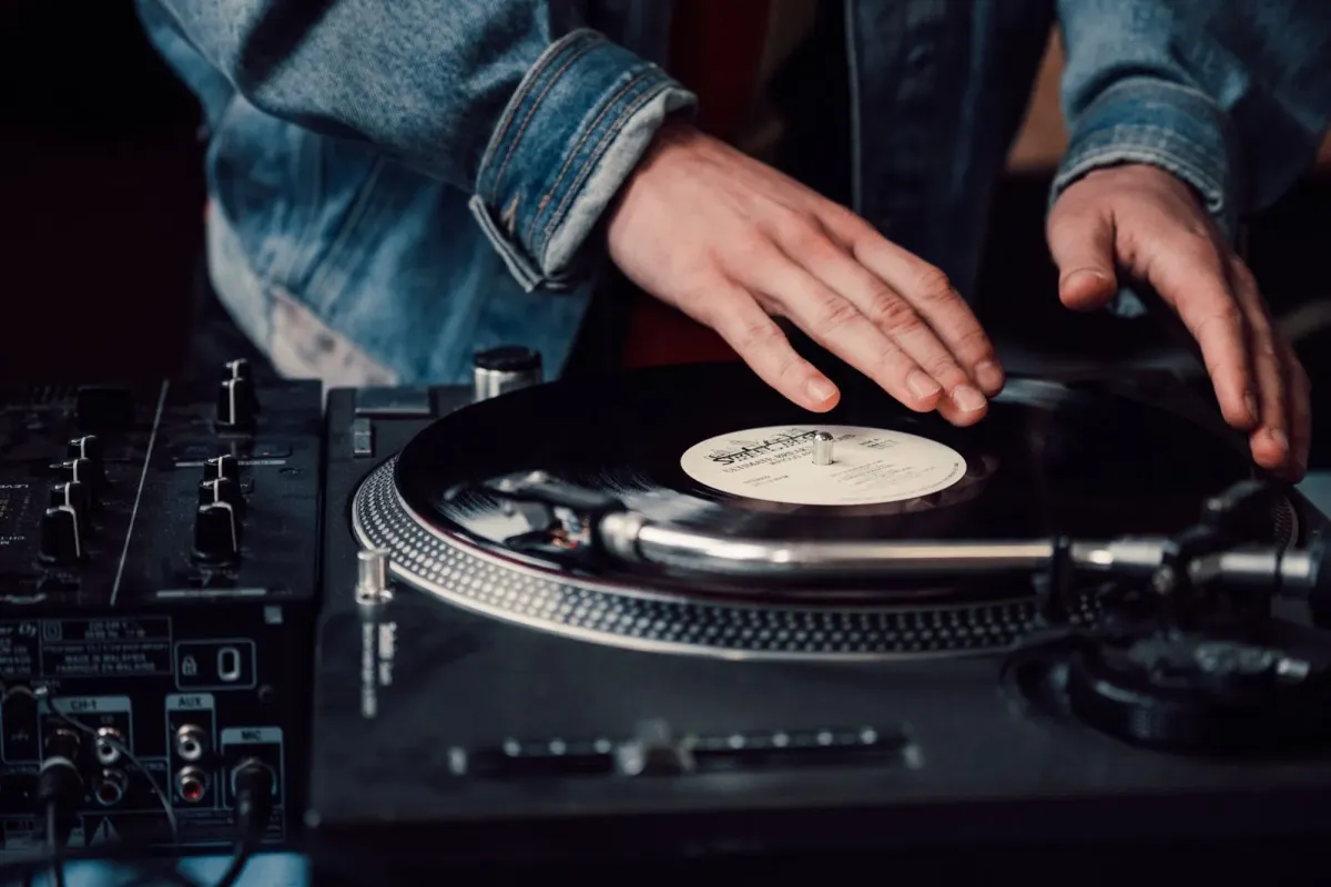 Mãos de um DJ sobre um disco de vinil em toca-discos, ao lado de mesa de som