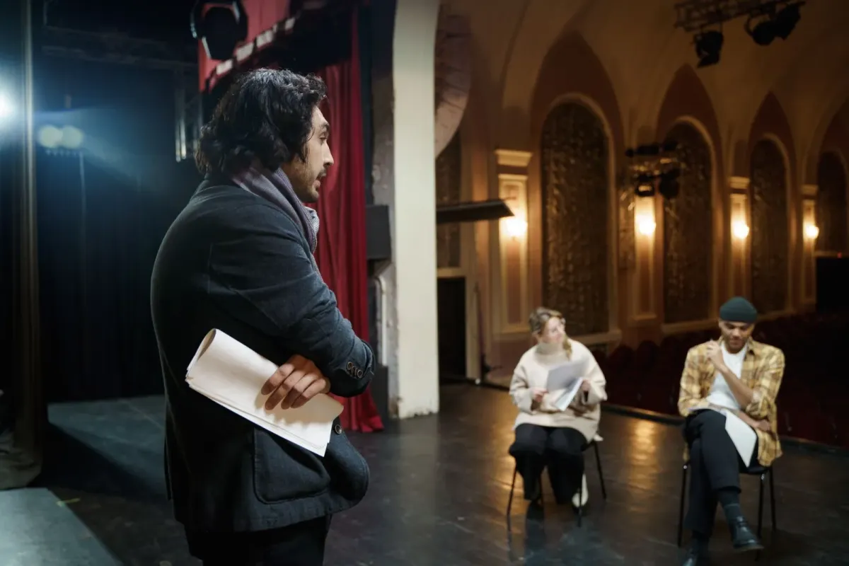 Homem de pé com roteiro sob o braço e dois atores sentados no palco de um teatro, em foto ilustrativa de ensaio