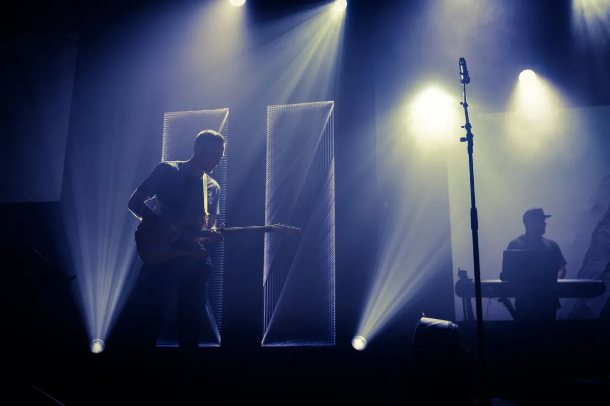 Guitarrista em silhueta no palco, com feixes de luz azulada e microfone ao lado
