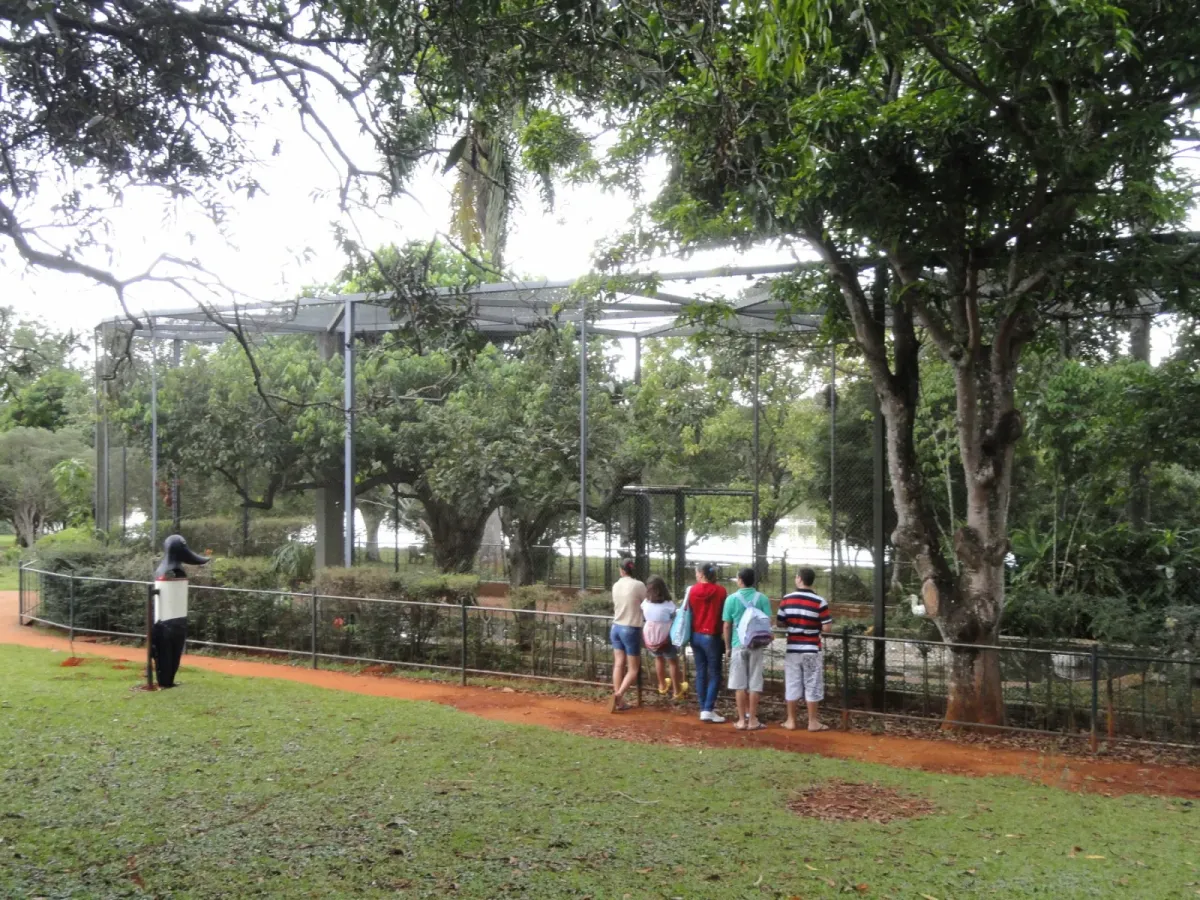Visitantes observam um recinto cercado entre árvores no Zoológico de Brasília, em caminho de terra vermelha