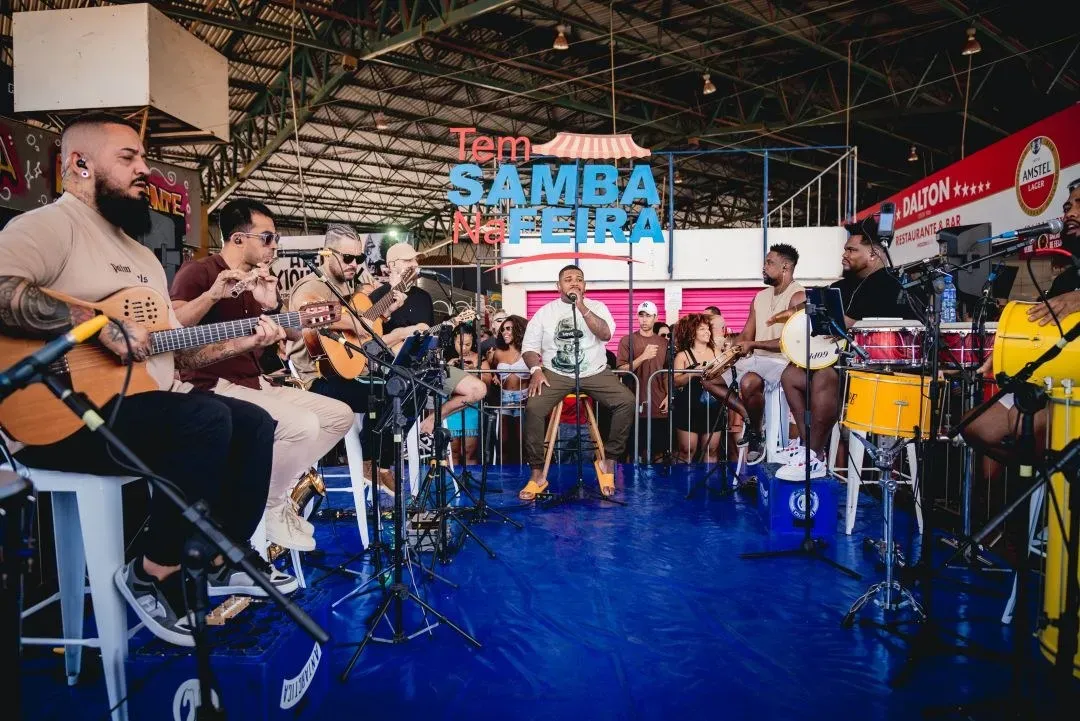 Roda de samba com violões, cavaquinho e percussão sob o telhado da Feira Permanente do Gama, com o letreiro Tem Samba na Feira